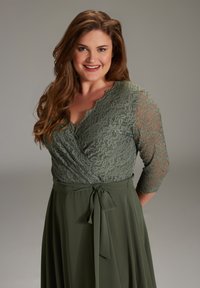 Grünes Kleid mit einem Spitzenoberteil und einem gewellten V-Ausschnitt, langen Spitzenärmeln und einem fließenden Rock, der an der Taille mit einem Stoffgürtel gebunden ist.