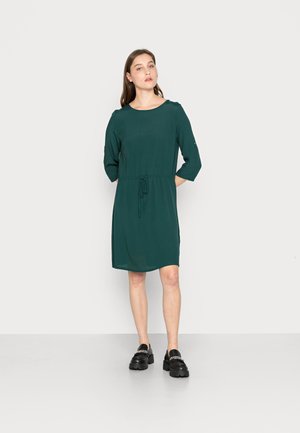 Vestido verde até ao joelho, com decote redondo, mangas 3/4 e detalhe de cintura ajustada por cordão. Combinado com sapatos pretos robustos.