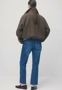 Osoba z długim kucykiem, ubrana w oversize'ową brązową kurtkę w kratę, niebieskie jeansy o prostych nogawkach, białe skarpetki i ciemnobrązowe mokasyny, stojąca na tle jednolitej ściany.