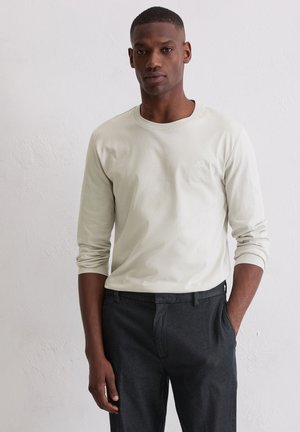 Helle beige Langarmbluse mit Rundhalsausschnitt, aus glattem Stoff; kombiniert mit dunkelgrauen Hosen. Dezentes Logo-Detail auf der Brust.