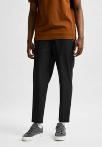 Svarta skräddarsydda byxor med smal passform, tillverkade av glatt tyg. Kombination med en brun t-shirt och grå sneakers, mot en neutral bakgrund.