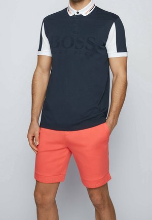 Mann trägt ein schwarz-weißes kurzärmeliges Poloshirt mit dem Text "BOSS" und helle Korallshorts mit dem Text "BOSS" am Saum.
