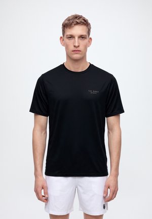 Un homme porte un T-shirt noir Ted Baker Sport et un short blanc sur un fond clair uni.