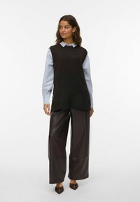 Oversized zwarte mouwloze trui over een lichtblauw overhemd, gecombineerd met brede bruine leren broeken en puntige schoenen.
