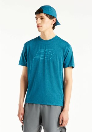Giovane uomo che indossa una t-shirt blu New Balance, pantaloncini grigi e un berretto blu portato al contrario, in piedi davanti a uno sfondo bianco.