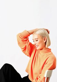 Maglione corto in tessuto arancione testurizzato con collo alto e motivo a coste, abbinato a pantaloni neri. Contrasto di colori vivaci e caldi.