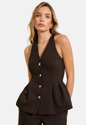 Frau trägt ärmelloses, schwarzes Peplum-Top mit goldenen Knöpfen und passende schwarze Hose, steht vor einfarbigem Hintergrund.