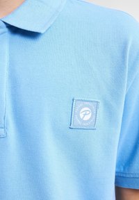 Světle modrá polo košile s texturovanou tkaninou, s vyšitým logem bílé barvy na levé straně hrudi. Jednobarevná s krátkými rukávy.