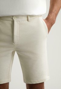 Pantaloni scurți de un bej deschis, dintr-un material texturat, având buzunare laterale, închidere cu buton și un design croit, ajungând până la mijlocul coapsei.
