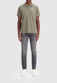 Olijfgroene polo met een kraag en een minimaal logo op de borst, gecombineerd met grijze denim jeans en witte sneakers.