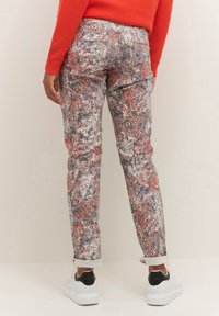 Person iført bukser med multikolor blomstermønster, rød langærmet top og hvide sneakers med sorte hælkonstruktioner, der står på en lys baggrund.