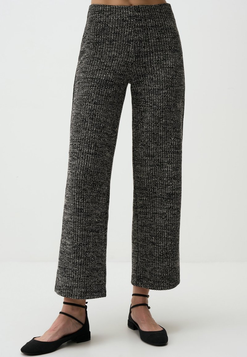 Femme portant un pantalon en maille gris foncé taille haute à jambes larges et des chaussures plates noires à bride à la cheville, debout sur un sol blanc.