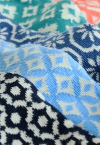 Tejido de punto en tonos de azul, verde azulado y marino, con patrones y texturas geométricas y acentos en blanco, doblado para su exhibición.