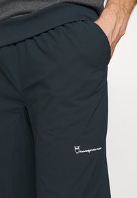 Tummansiniset urheiluhousut, joissa on joustava vyötärönauha ja sivutaskut. Reisiosassa valkoinen Knowledge Cotton Apparel -logo.