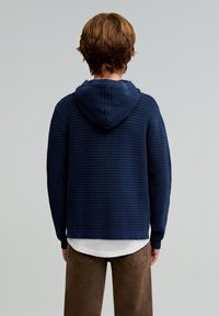 Granatowy, dziany hoodie z teksturowanym wzorem i przednią kieszenią. Zestawiony z brązowymi spodniami, pokazany z tyłu na jasnym tle.
