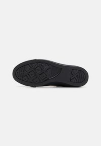 Semelle de sneaker en caoutchouc noir présentant un motif de grip texturé avec des formes géométriques et un bord lisse. Logo visible sur le côté.