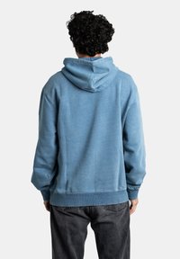 Sudadera azul con capucha ajustable, puños y bajo acanalados, hecha de una tela suave, mostrada desde la parte trasera con un ajuste relajado sobre unos jeans oscuros.