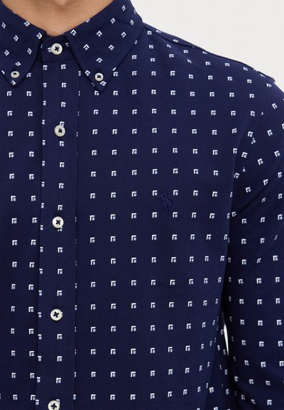Tamsiai mėlyna marškiniai su užsegimu, papuošti mažų baltų kvadratėlių raštu, su mygtukų apykakle ir subtiliu logotipu ant krūtinės.