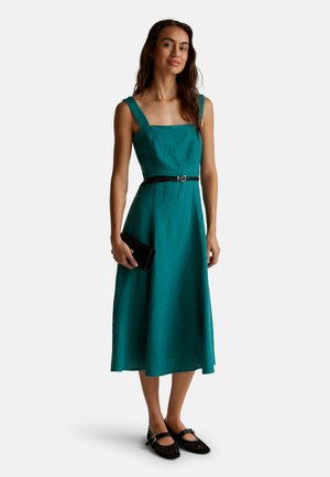 Abito di lino teal con scollo quadrato, spalline larghe e vita regolata da una cintura. Abbinato a una pochette nera e scarpe in rete nere.
