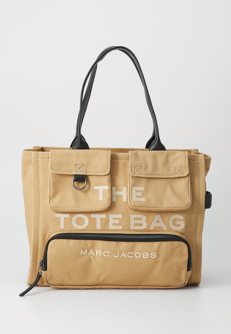 Canvas draagtas in bruin met zwarte leren handvatten. Bevat voorzakken en een gevlokt compartiment, plus gedurfde tekst: "DE DRAAGTAS" en "MARC JACOBS."