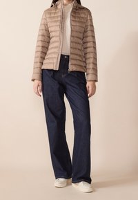 Beżowa pikowana kurtka z poziomym przeszyciem, zamkiem na przodzie i kapturem, zestawiona z ciemnoniebieskimi szerokimi dżinsami i białymi tenisówkami.