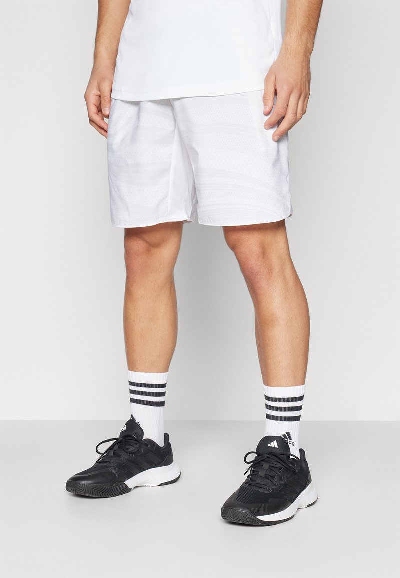 Weiße Sportshorts mit einem strukturierten Muster, kombiniert mit schwarzen Sneakers und gestreiften weißen Socken. Fokus auf klare Linien und sportlichem Look.