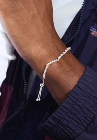 Pulsera de cuentas de plata con cierre deslizante, que cuenta con dos pequeñas cuentas colgantes. Lleva puesta en una muñeca de tono de piel oscura con una apariencia texturizada.
