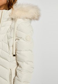 Lys beige pufferjakke med hætte i faux fur, diamant quiltet mønster og et logo patch på ærmet. Har en lynlåslukning.