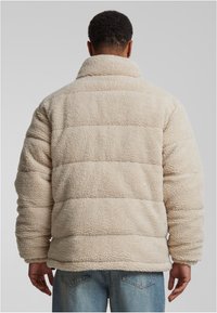 Beige fleecetröja med hög krage, texturerad yta, kviltat mönster och långa ärmar, visad från baksidan.