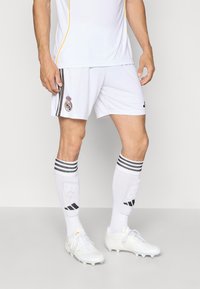 Maglia da calcio bianca con accenti gialli, abbinata a pantaloni corti bianchi con un emblema di squadra. I calzini bianchi hanno strisce nere e logo. Le scarpe sono bianche.