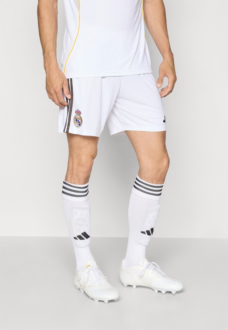 Maglia da calcio bianca con accenti gialli, abbinata a pantaloni corti bianchi con un emblema di squadra. I calzini bianchi hanno strisce nere e logo. Le scarpe sono bianche.