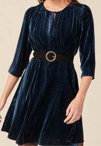 Vestido de terciopelo azul marino oscuro con mangas tres cuartos, escote redondeado y un corte en forma de llave, ceñido con un cinturón negro que presenta una hebilla redonda de plata.