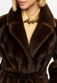 Brun faux fur frakke med en stor krave, bælte i taljen og en blød tekstur. Båret over en guld knap-op skjorte og en kraftig guld halskæde.