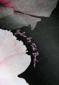 Un textile présentant un motif floral dans des nuances de rose et de blanc, avec un fond texturé noir. "Conçu par Ted Baker" est imprimé en cursive rose.