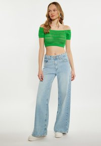 Crop top verde, cu umerii la vedere, tricotez, asortat cu blugi largi de culoare albastru deschis. Modelul poartă adidași albi, prezentat pe un fundal neutru.