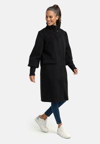 Zwarte wolmix jas met hoge kraag, lange mouwen en voorzak. Draagt over een blauwe jeans en witte sneakers. Soepele textuur.