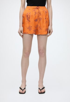 Persoană purtând pantaloni scurți portocalii cu imprimeu floral și șnur în talie, top negru și sandale negre cu vârful deschis, stând pe un fundal alb.