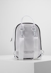 Malý bílý batoh s nastavitelnými bílými popruhy a černým lemováním, umístěný na bílém podstavci na pozadí z čistě bílé barvy.