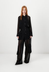 Crna prozirna košulja s dugim rukavima i dugmadi, uparena s crnim hlačama viskog struka koje imaju cargo džepove i širok kroj.