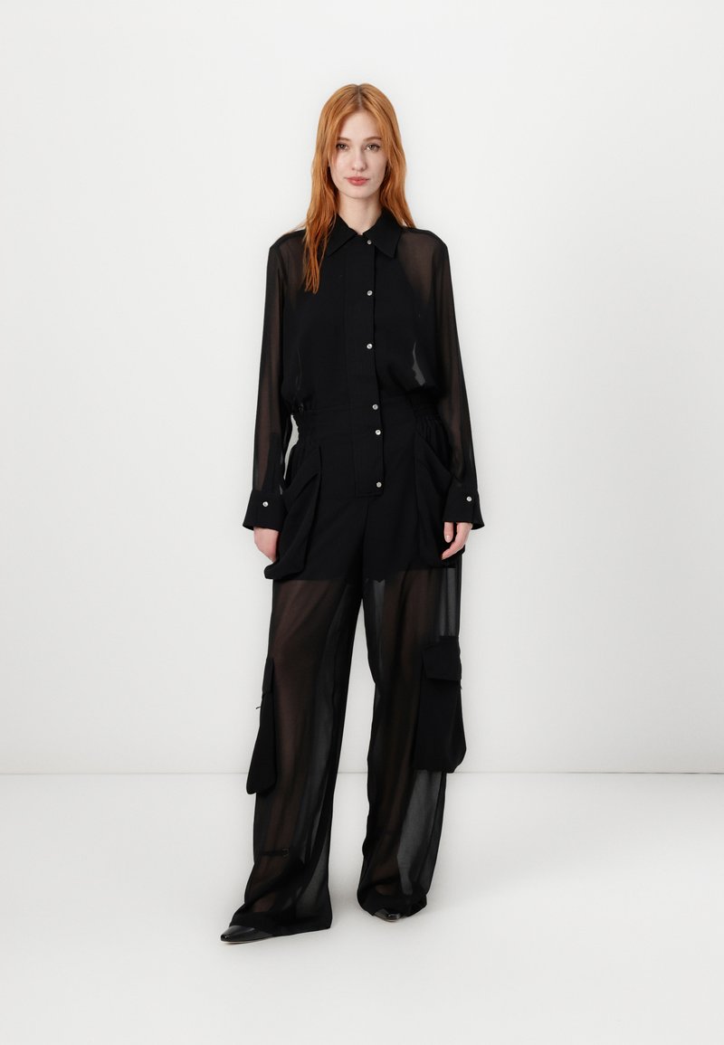 Crna prozirna košulja s dugim rukavima i dugmadi, uparena s crnim hlačama viskog struka koje imaju cargo džepove i širok kroj.