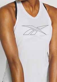 Vit sporttopp med rund halsringning. Har en grå Reebok-logotyp på bröstet. Slät tyg, slim fit med svarta band på baksidan.