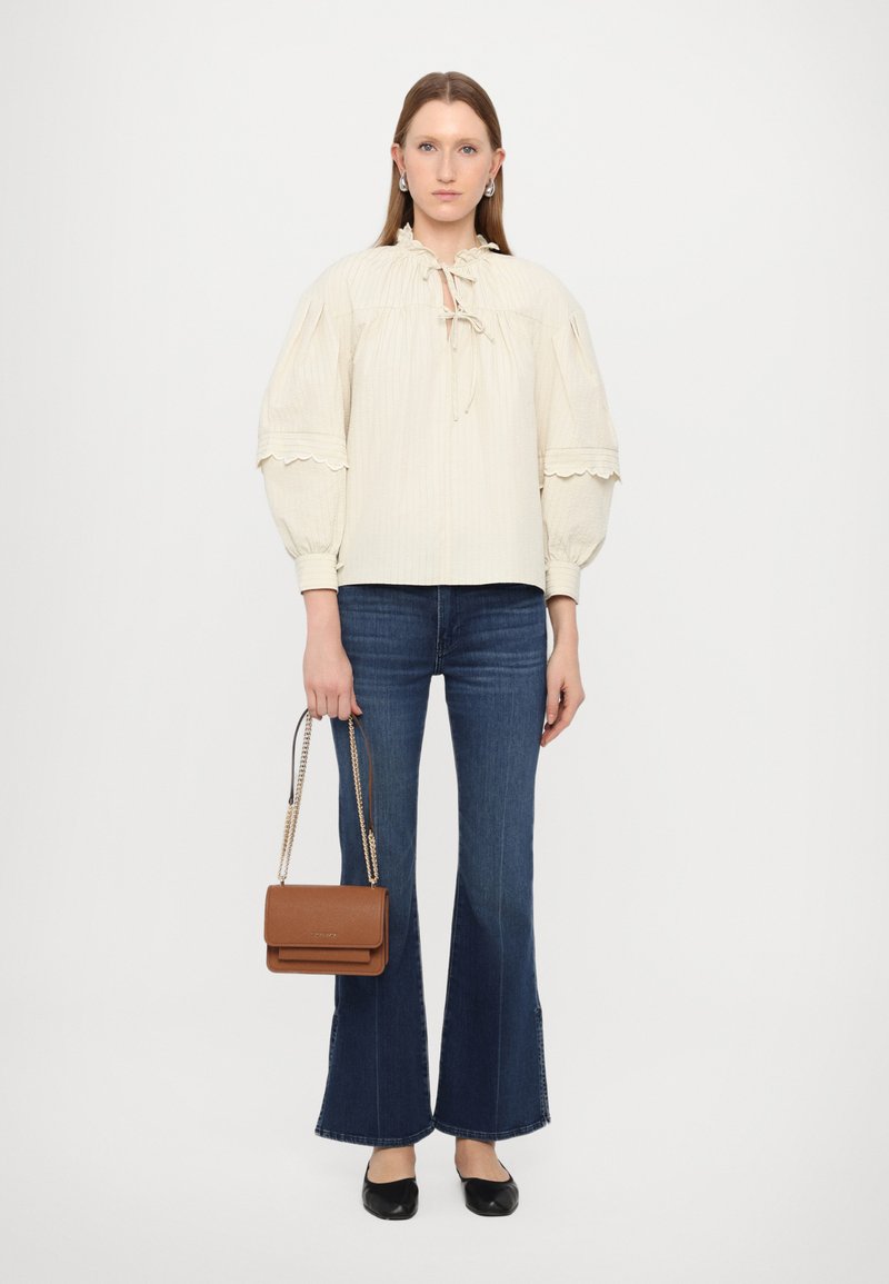 Blusa cremoso, con texture, maniche a sbuffo e collo con fiocco, abbinata a jeans blu a palazzo e a una piccola borsa a tracolla marrone. Ballerine nere.