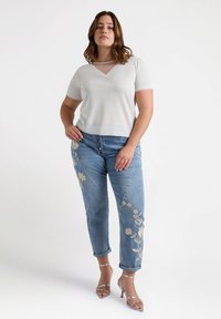 Jasnoszara bluzka z krótkim rękawem, dekoltem w serek i przezroczystymi detalami, połączona z niebieskimi dżinsami z beżowym haftem kwiatowym oraz srebrnymi sandałami na wysokim obcasie.