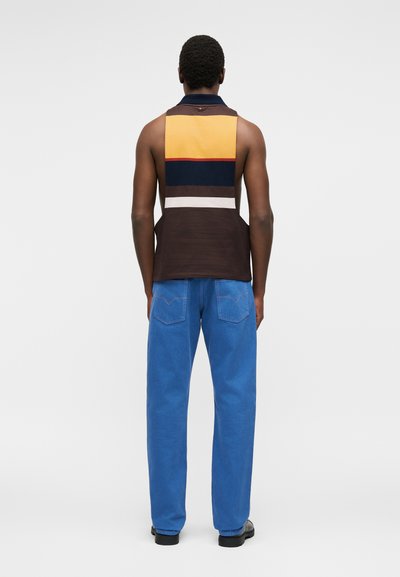 Άνδρας που φοράει αμάνικη μπλούζα με χρωματικούς συνδυασμούς σε κίτρινο, ναυτικό μπλε, καφέ και λευκές ρίγες, μπλε τζιν και μαύρα παπούτσια, γυρισμένος με την πλάτη σε λευκό φόντο.