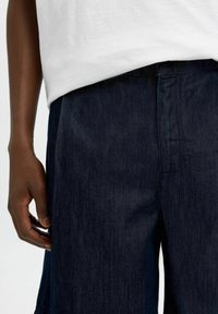 Mörkblå denimbyxor med rak bendesign, som har en strukturerad yta och en synlig midja. Tyget har en lätt lyster.