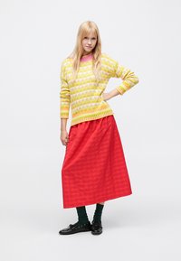 Žltý pletený sveter s kvetinovým vzorom, skombinovaný s dlhou červenou sukňou s pásikmi. Nosený so zelenými ponožkami a čiernou obuvou.