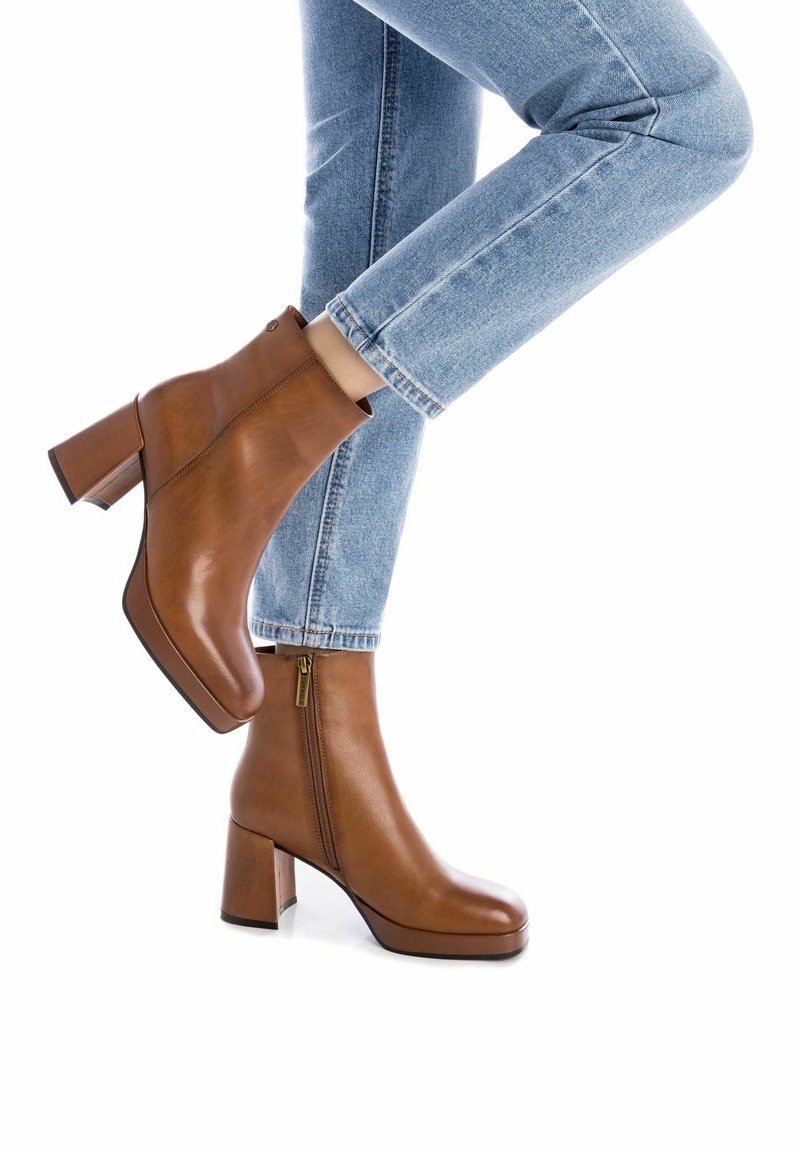 Botas de cuero marrón hasta el tobillo con un tacón cuadrado, que presentan una cremallera lateral y una textura suave, llevadas con unos vaqueros de tela vaquera azul claro remangados.
