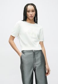 Femeie cu cozi împletite lungi, purtând un pulover alb cu mâneci scurte și pantaloni gri cu talie înaltă, pozând cu o mână în buzunar.