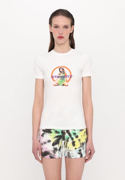 Witte nauwsluitende t-shirt met een kleurrijke grafische print van een zittende figuur en de tekst "STIEGLITZ 1988." Gecombineerd met tie-dye shorts in roze, groen en zwart.