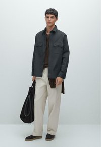 Dark gray button-up jacket with two chest pockets, layered over a brown shirt. Cream wide-leg trousers and a dark tote bag.